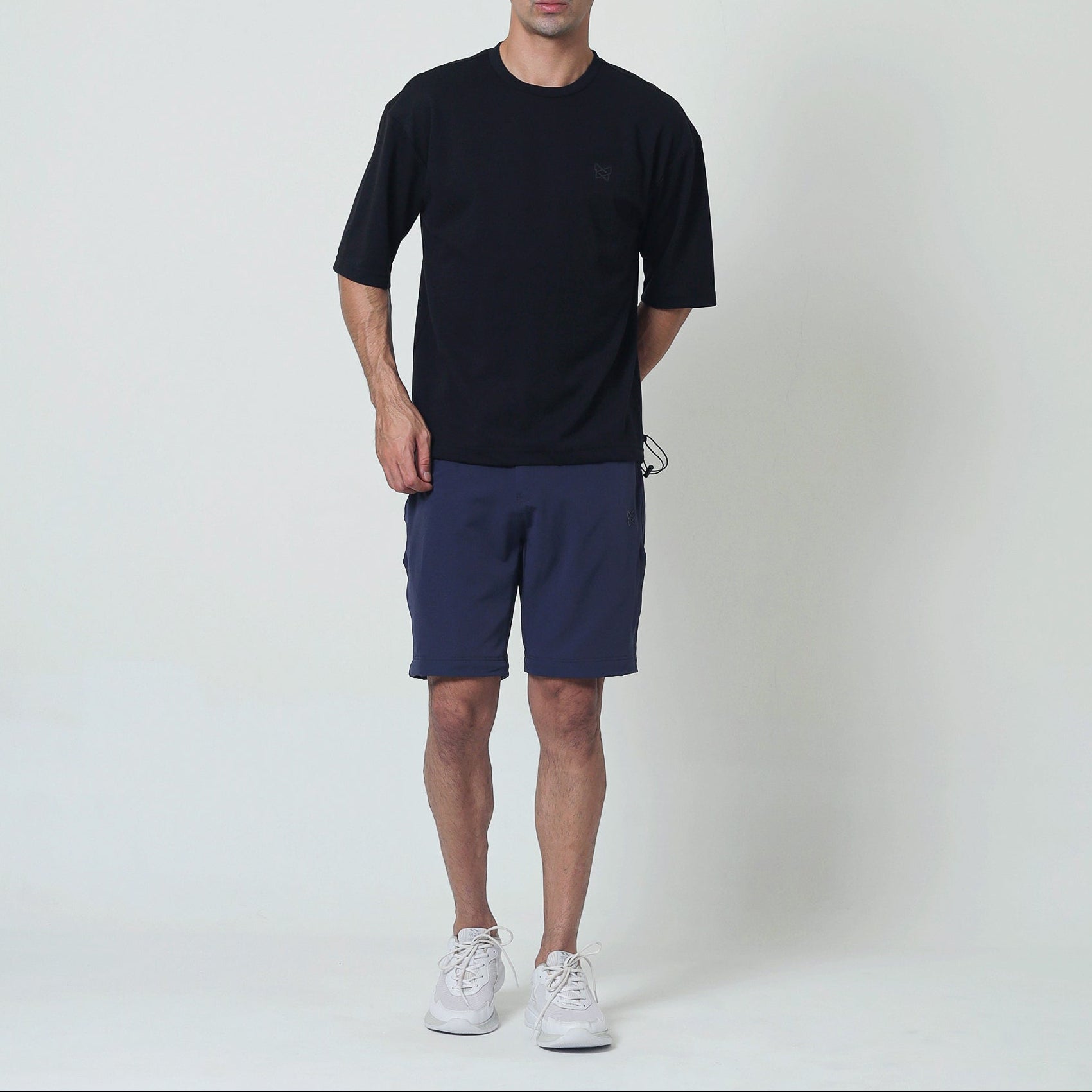 A man modeling a black short-sleeve crewneck t-shirt and navy blue athletic shorts, paired with white sneakers, standing against a light gray studio background.